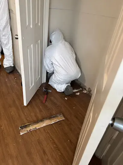 Technician in a protective suit tearing out wet baseboard and flooring at a doorway